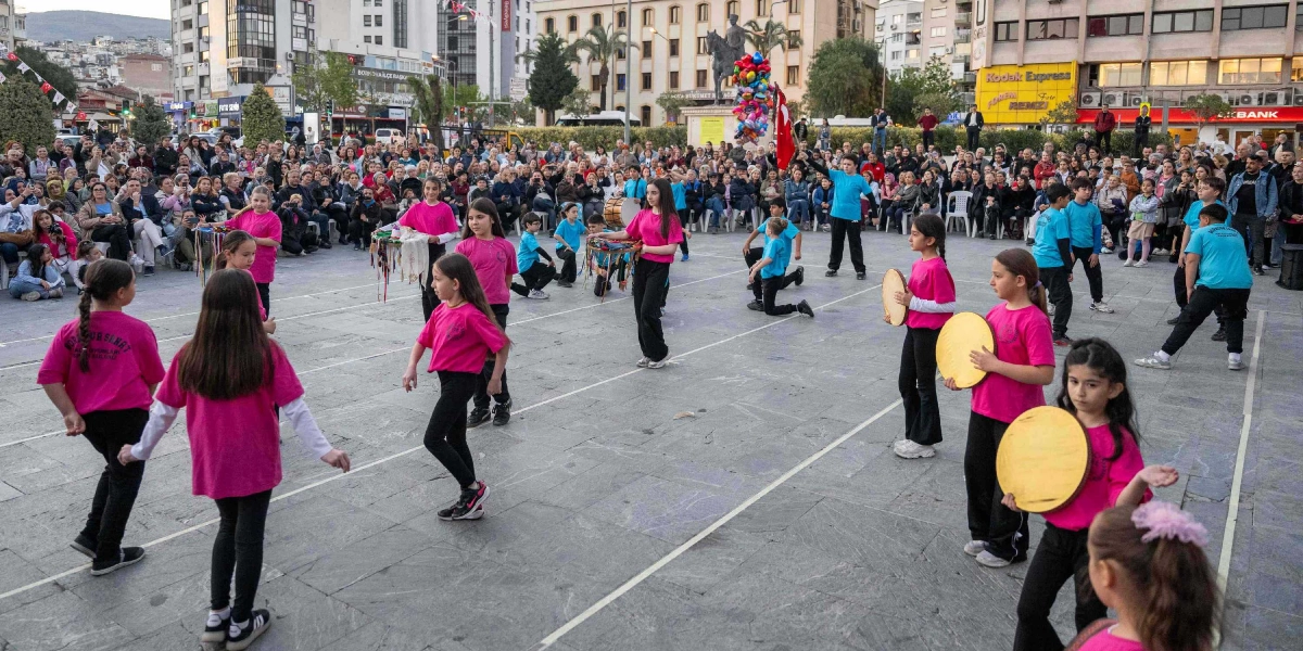 Bornova Cumhuriyet Meydanı'nda düzenlenen Dünya Dans Günü kutlamasında, pembe tişörtlü kız çocukları ve mavi tişörtlü erkek çocuklarından oluşan bir grup, ellerinde teflerle halk dansları performansı sergiliyor. Meydanın çevresinde toplanan kalabalık bir izleyici grubu gösteriyi izlerken, arka planda Atatürk heykeli, çevredeki binalar ve balon tutan bir kişi görülüyor.