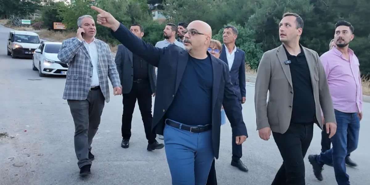 Bornova İzyuva bölgesinde incelemelerde bulunan bir grup yetkili, asfalt bir yol üzerinde yürürken görülüyor. Grubun önünde yer alan gözlüklü bir erkek, eliyle ilerideki bir noktayı işaret ederek açıklama yapıyor. Yanındaki diğer kişiler dikkatle onu dinliyor. Arka planda ağaçlık bir alan ve yol kenarında park halindeki araçlar bulunuyor.
