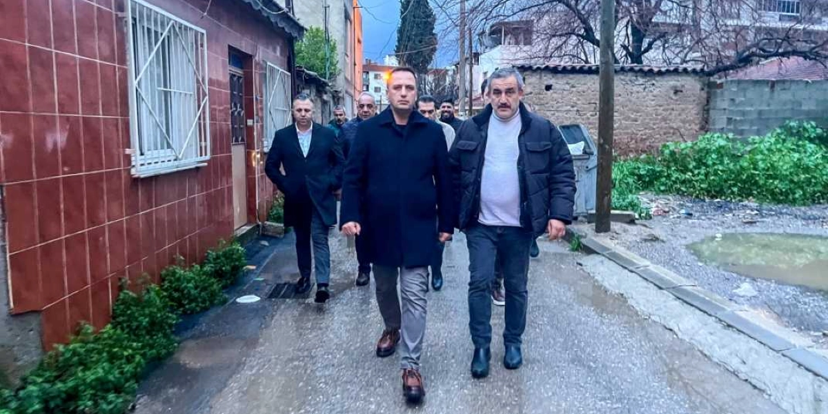 Bornova Belediye Başkanı Mustafa İduğ ve beraberindeki heyet, Doğanlar mahallesindeki bir sokakta incelemelerde bulunuyor. Koyu renkli kabanlar giymiş olan grup, yan yana yürüyerek çevreyi gözlemliyor. Sol tarafta kahverengi fayans kaplı bir bina, sağ tarafta ise taş duvarlı bir bahçe ve boş bir arazi yer alıyor.