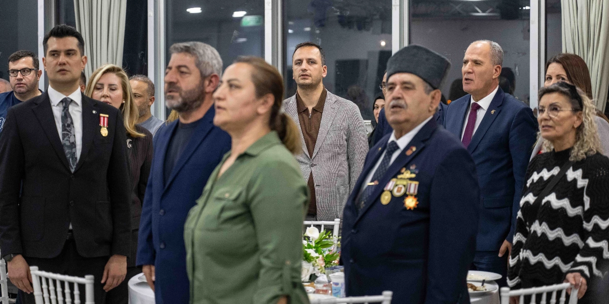 Kapalı bir mekanda, resmi bir etkinlikte ayakta duran ve ciddiyetle bir noktaya bakan bir grup insan. Ön planda, üzerinde madalyalar bulunan lacivert takım elbiseli ve kalpaklı bir beyefendi ile yanında haki renkli bir ceket giyen bir kadın yer alıyor. Arka planda ise takım elbiseli erkekler ve farklı kıyafetler içindeki kadınlar bulunuyor. Masaların üzerinde beyaz örtüler ve çiçek aranjmanları görülüyor.