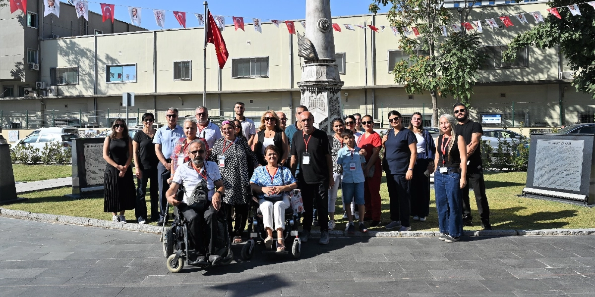 Açık havada, üzerinde Türk bayrakları ve Atatürk posterlerinin asılı olduğu süslemelerin bulunduğu bir meydanda, bir anıtın önünde bir araya gelmiş çeşitli yaş ve gruplardan oluşan kalabalık bir grup insan. Grubun ön sırasında tekerlekli sandalyede oturan iki kişi ve yanlarında ayakta duran katılımcılar yer alıyor. Arka planda ise ağaçlar, park alanı ve üzerinde yazılı metinler bulunan iki adet anıt panosu görülüyor.