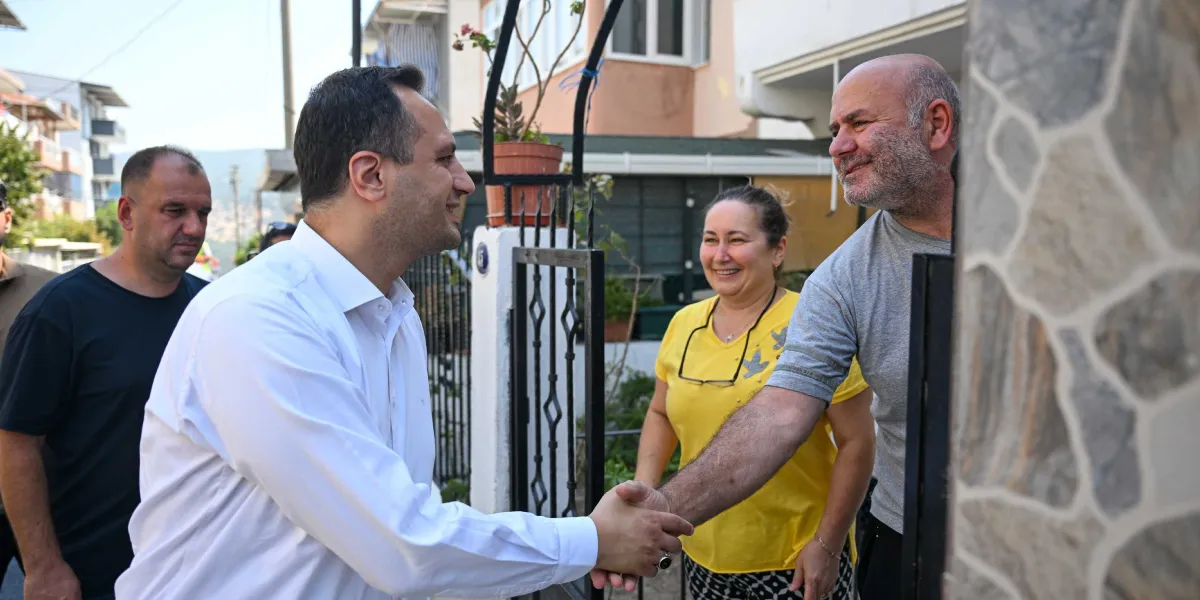 Beyaz gömlekli bir erkek, bir evin bahçe kapısında sakallı bir adamla tokalaşırken, yanlarında duran sarı tişörtlü bir kadın gülümseyerek onları izliyor. Arka planda ise başka bir erkek daha bulunuyor.