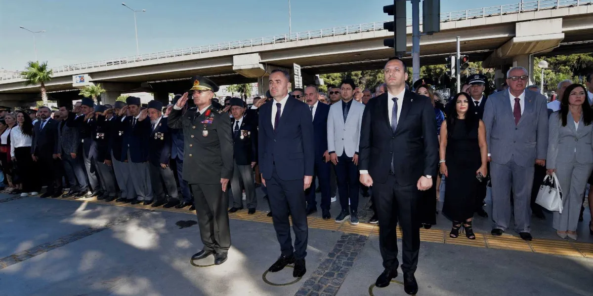 Açık havada, bir üst geçidin altında düzenlenen resmi törende, aralarında askeri üniformalı bir subay, takım elbiseli yetkililer ve gazilerin bulunduğu bir grup insan saygı duruşunda bulunuyor. Askeri üniformalı subay selam verirken, diğer katılımcılar elleri yanlarında hazır olda bekliyor. Arka planda trafik ışıkları ve beton bir viyadük yapısı görülüyor.