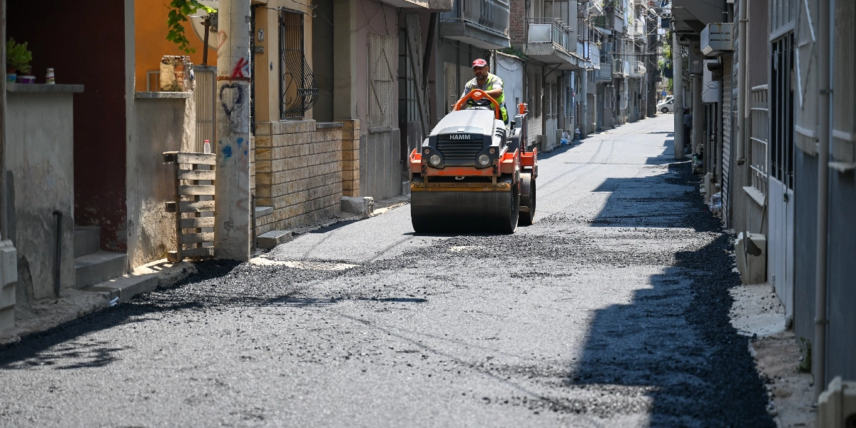 Çamdibi'nde dar bir sokakta, yeni dökülmüş asfaltı düzleştiren operatör yönetimindeki turuncu renkli bir Hamm silindir iş makinesi ilerliyor.