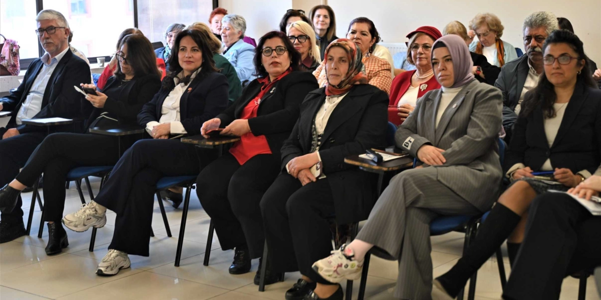 Bir toplantı salonunda yan yana dizilmiş sandalyelerde oturan, çoğunluğu kadınlardan oluşan bir grup insan dikkatle karşıya bakıyor. Katılımcıların bir kısmı resmi kıyafetler, ceketler ve gömlekler giymiş durumda; aralarında başörtülü kadınlar da bulunuyor. En ön sırada oturanlar arasında gri çizgili takım elbiseli, başörtülü bir kadın ve yanında siyah ceketli, eşarplı bir kadın yer alıyor. Arka sıralarda ise farklı yaş gruplarından ve giyim tarzlarından katılımcılar bulunuyor. Salonun genel atmosferi ciddi ve odaklanmış bir toplantı ortamını yansıtıyor.