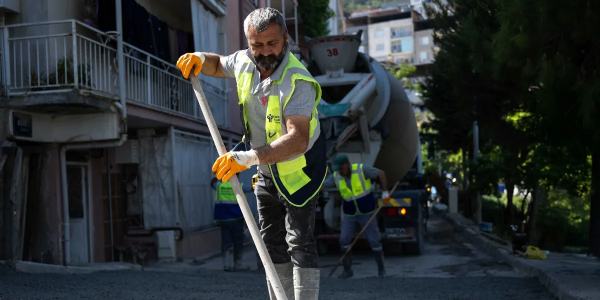 Bornova Belediyesi yeleği giymiş bir işçi, yokuşlu bir sokakta kürekle beton düzeltme çalışması yapıyor. Arka planda beton mikseri ve diğer belediye işçileri yol çalışmasına devam ediyor.