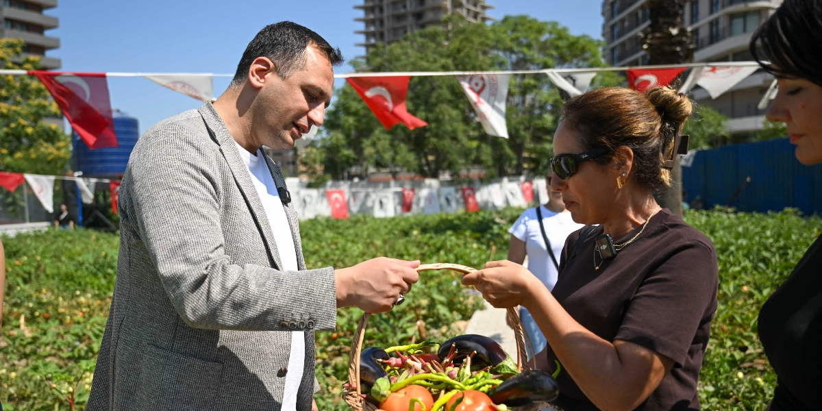 Gri ceketli bir erkek ve güneş gözlüklü bir kadın, açık havada bulunan bir sebze bahçesinde ellerinde içinde patlıcan, domates ve biber bulunan hasır bir sepeti birlikte tutuyor. Arka planda Türk bayraklarıyla süslenmiş bir alan ve yüksek binalar görülüyor.