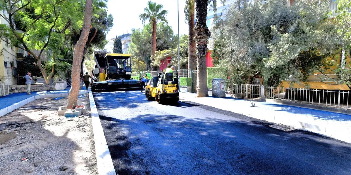 Ağaçlıklı bir sokakta devam eden yol yenileme çalışmaları kapsamında, iş makineleri yeni dökülmüş asfalt üzerinde ilerliyor. Sarı renkli bir silindir ve arkasında asfalt serme makinesi çalışırken, yolun kenarlarında mavi renge boyanmış kaldırımlar ve çevredeki ağaçlar görülüyor. Çalışan işçiler ve sokak boyunca uzanan binalar arka planda yer alıyor.
