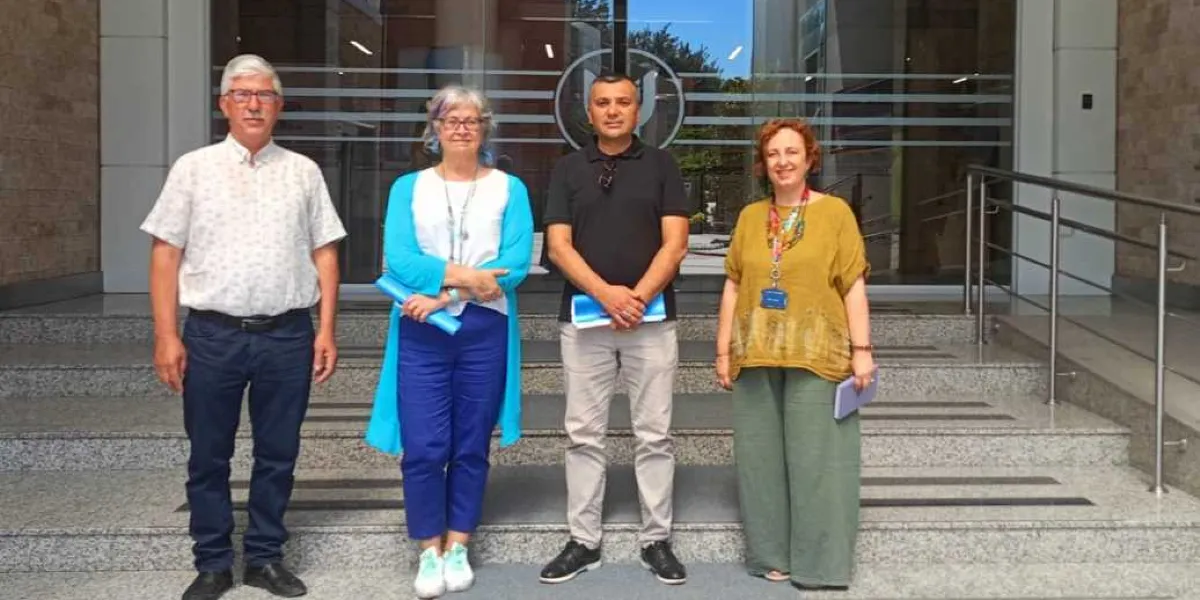 Modern bir binanın girişindeki merdivenlerde yan yana duran, ikisi kadın ikisi erkek dört kişi. Soldan sağa doğru; beyaz kısa kollu gömlek ve lacivert pantolon giyen gözlüklü bir erkek, beyaz bluz üzerine mavi hırka ve mavi pantolon giyen gri saçlı bir kadın, siyah polo yaka tişört ve bej pantolon giyen bir erkek ve hardal sarısı bluz ile yeşil bol pantolon giyen, boynunda yaka kartı asılı bir kadın yer alıyor. Kişilerin ellerinde mavi dosyalar ve not defterleri bulunuyor.