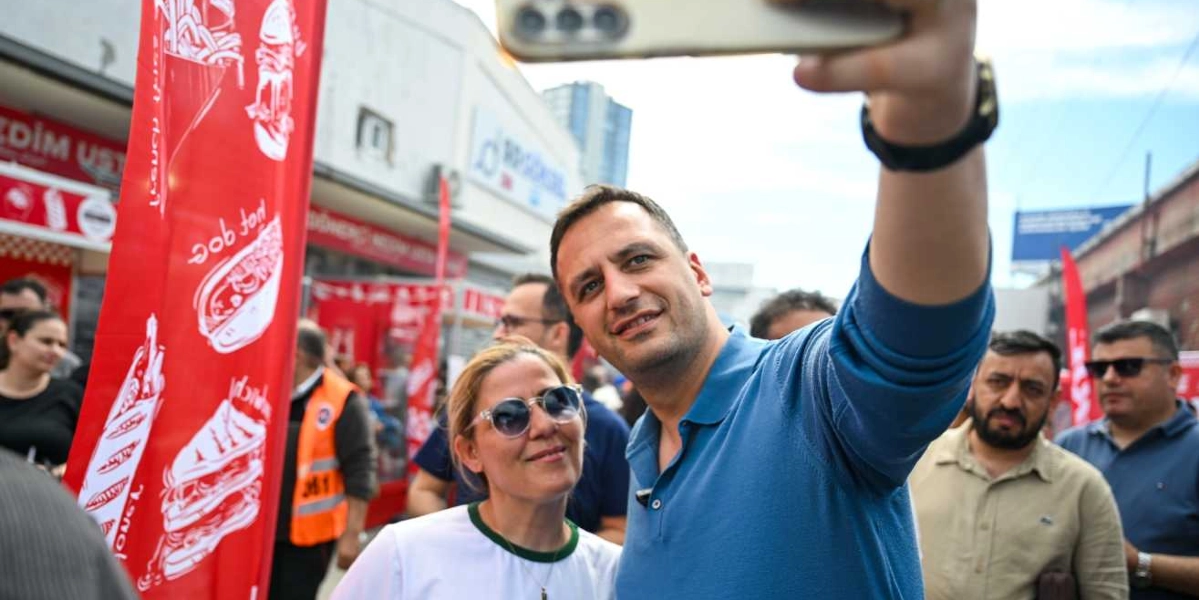 Mavi polo yaka tişörtlü bir erkek, yanında duran güneş gözlüklü bir kadınla birlikte akıllı telefonuyla selfie çekiyor. Arka planda Bornova'daki sanayi bölgesinde kurulan festival alanında kırmızı renkli yiyecek standı flamaları ve kalabalık bir grup yer alıyor.