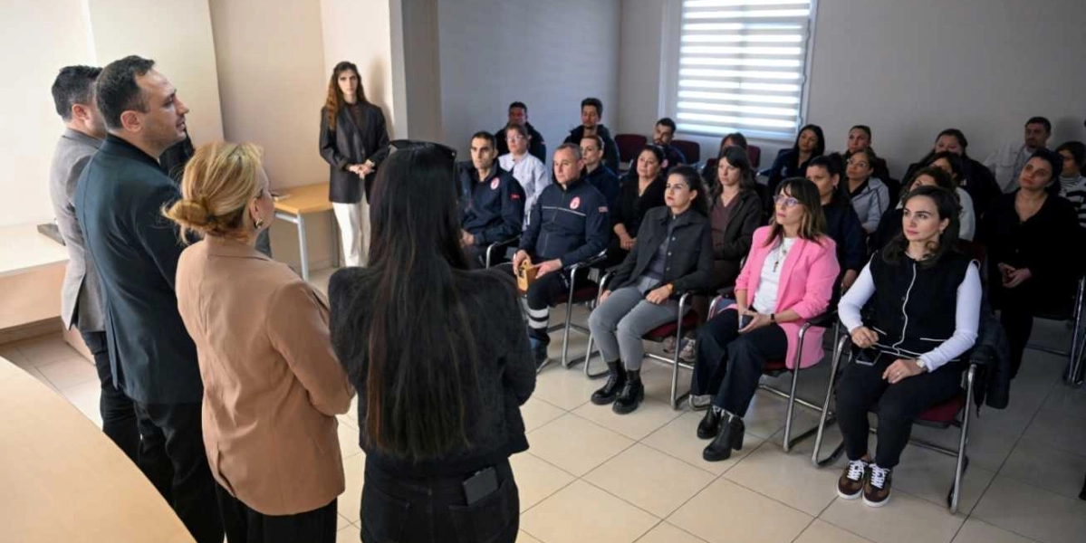Kapalı bir toplantı salonunda, ayakta duran birkaç kişinin bir grup katılımcıya sunum yaptığı veya bilgilendirme toplantısı gerçekleştirdiği an. Ön planda sırtları dönük şekilde duran üç kişi, karşılarında sandalyelerde oturan ve dikkatle dinleyen yaklaşık yirmi kişilik bir topluluğa hitap ediyor. Katılımcıların çoğu kadınlardan oluşuyor ve ortamda resmi bir atmosfer hakim.