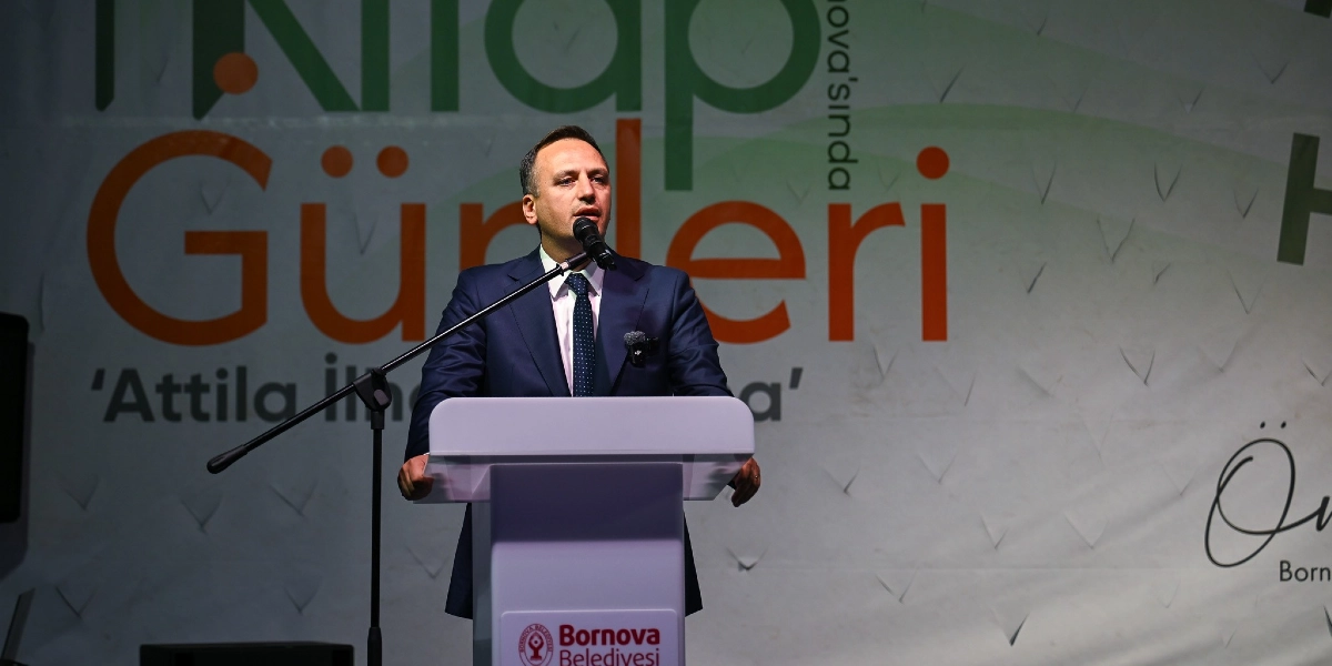 Koyu renk takım elbiseli ve kravatlı bir erkek, üzerinde Bornova Belediyesi logosu bulunan beyaz bir kürsüde mikrofon başında konuşma yapıyor. Arka planda 