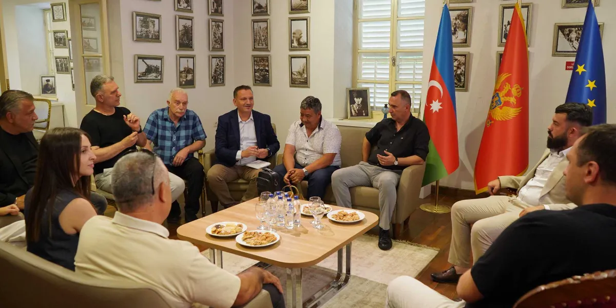 Bornova Belediyesi yetkilileri ve Makedon heyet üyeleri, bir toplantı odasında karşılıklı koltuklarda oturarak sohbet ediyor. Ortadaki ahşap masanın üzerinde ikramlık tabaklar ve su şişeleri bulunuyor. Arka planda duvarda çerçeveli fotoğraflar asılıyken, sağ tarafta Azerbaycan, Karadağ ve Avrupa Birliği bayrakları yan yana duruyor.