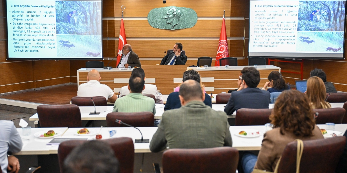 Belediye meclis salonunda düzenlenen bir toplantıda, katılımcılar önlerindeki masalarda otururken karşılarındaki iki büyük ekranda Bornova'daki biyoçeşitlilik envanter çalışmalarıyla ilgili sunum yansıtılıyor. Ekranda, 7 öğretim görevlisiyle 54 saha çalışması yapıldığı, 433 bitki, 23 sürüngen, 22 memeli ve 41 kuş türünün kayıt altına alındığı bilgisi yer alıyor; ayrıca sahada çalışma yapan bir araştırmacı ve koşan bir yaban hayvanının görselleri bulunuyor. Salonun ön tarafındaki kürsüde iki yetkili otururken, arka planda duvarda Atatürk kabartması ve iki yanında Türk bayrağı ile Bornova Belediyesi'nin flaması yer alıyor.