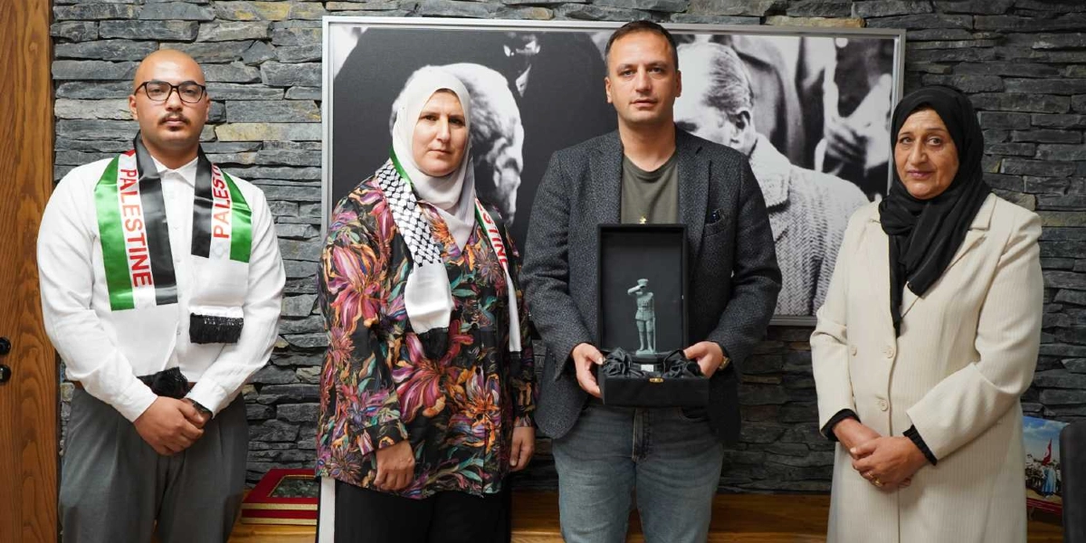 Taş duvarlı bir arka plan önünde yan yana duran dört kişi; soldan sağa doğru, boynunda Filistin bayraklı bir atkı bulunan gözlüklü bir erkek, üzerinde Filistin kefiyesi olan başörtülü bir kadın, elinde içinde asker figürü bulunan bir hediye kutusu tutan gri ceketli bir erkek ve siyah başörtülü bir kadın yer alıyor. Arka planda Mustafa Kemal Atatürk'ün yer aldığı siyah beyaz bir tablo bulunuyor.
