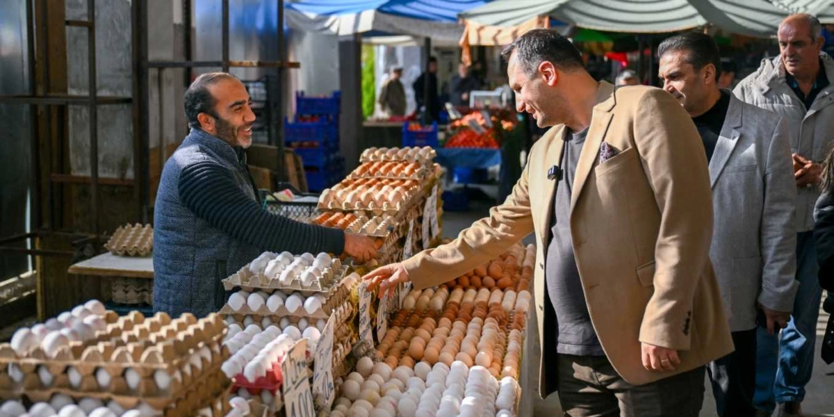 Bej renkli ceket giyen bir yetkili, pazar tezgahındaki yumurtaları incelerken tezgah sahibi esnafla sohbet ediyor. Arka planda diğer pazar esnafları ve alışveriş yapan kişiler yer alıyor.