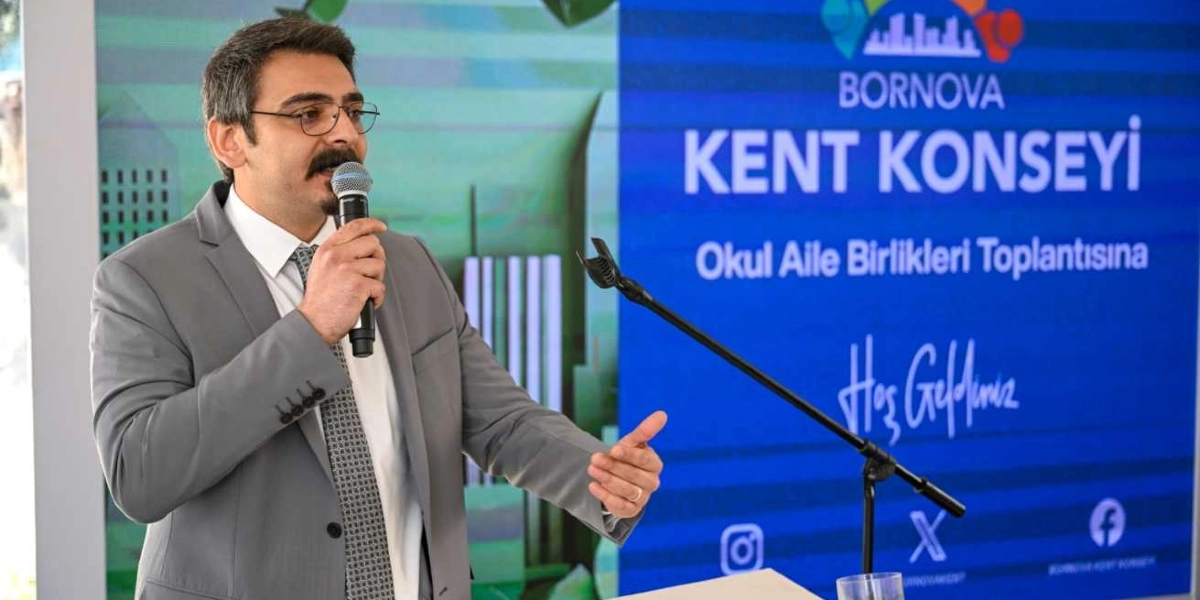 Gözlüklü, bıyıklı ve gri takım elbiseli bir erkek, Bornova Kent Konseyi Okul Aile Birlikleri Toplantısı'nda mikrofonla konuşma yapıyor. Arka planda üzerinde Bornova Kent Konseyi logosunun, 