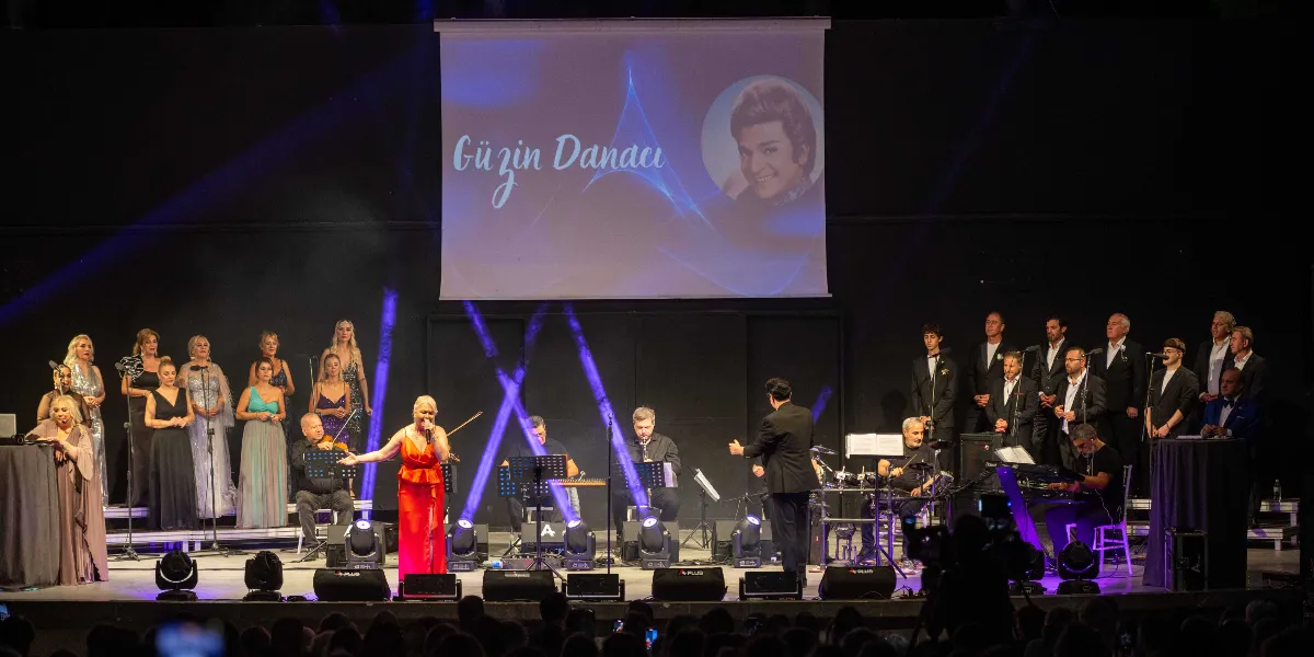 Sahne üzerinde kırmızı elbiseli bir kadın solist mikrofonla şarkı söylerken, arkasında orkestra ve koro üyeleri yer alıyor. Sahnenin arka planındaki büyük ekranda, üzerinde 