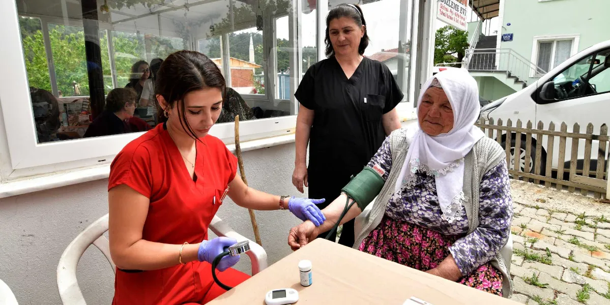 Kırmızı üniformalı genç bir sağlık çalışanı, dış mekanda oturan yaşlı bir kadının tansiyonunu ölçüyor. Sağlık çalışanının yanında siyah üniformalı başka bir kadın görevli daha bulunuyor. Yaşlı kadın başında beyaz bir başörtüsüyle masada oturuyor, masanın üzerinde ise küçük bir tıbbi cihaz ve ilaç kutusu yer alıyor.