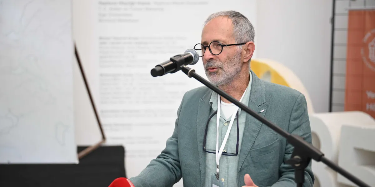 Gri ceketli, gözlüklü ve beyaz yakalı bir adam, bir etkinlikte mikrofon başında konuşma yapıyor. Boynunda bir yaka kartı asılı olan adamın arkasında üzerinde metinler bulunan beyaz bir pano ve sağ tarafta kısmen görünen turuncu bir panel yer alıyor.