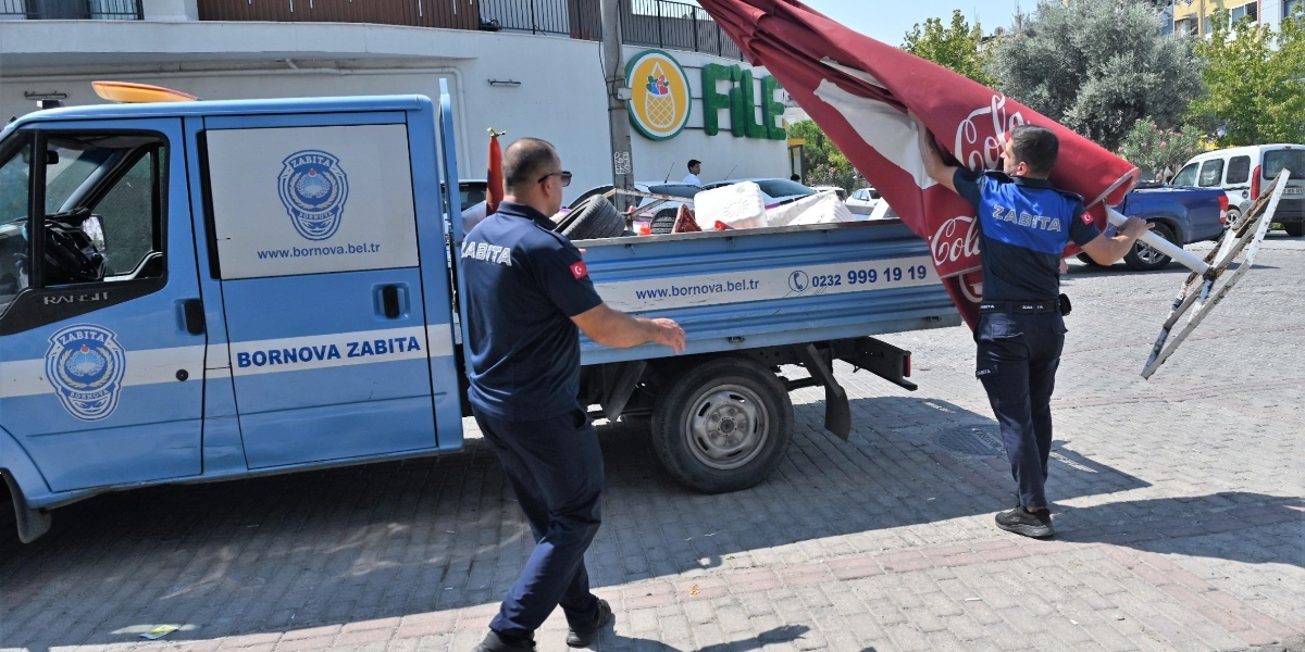 Bornova Belediyesi zabıta ekipleri, kaldırım işgali yapan büyük bir şemsiyeyi ve diğer malzemeleri toplayarak açık kasa zabıta kamyonetine yüklüyor.