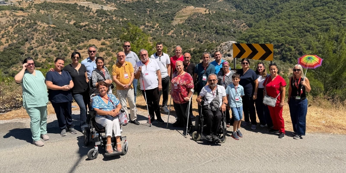Açık havada, dağlık ve ağaçlıklı bir arazinin önündeki asfalt yolda yan yana duran, aralarında tekerlekli sandalye kullananların ve görme engelli bireylerin de bulunduğu kalabalık bir grup insan. Grubun önünde tekerlekli sandalyede oturan bir kadın ve bir erkek bulunurken, diğer katılımcılar ayakta duruyor. Bazı kişiler ellerinde beyaz baston tutuyor, sağ taraftaki bir kadın ise güneşten korunmak için gökkuşağı renklerinde bir şemsiye taşıyor. Arka planda sarı zemin üzerine siyah ok işareti bulunan bir trafik levhası yer alıyor.