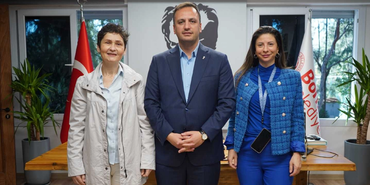 Bornova Belediye Başkanı Ömer Eşmi, yanında iki kadın yetkili ile birlikte ofis ortamında yan yana duruyor. Ortada lacivert takım elbiseli Ömer Eşmi, solunda açık renkli bir ceket giyen kısa saçlı bir kadın, sağında ise mavi bir ceket ve pantolon takımı giyen uzun saçlı bir kadın yer alıyor. Arka planda Türk bayrağı, Bornova Belediyesi'nin logosunun bulunduğu bir flama ve Atatürk silüeti görünüyor.