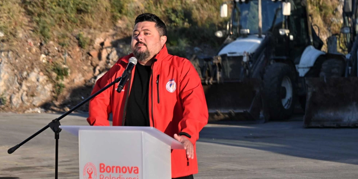 Kırmızı ceketli, sakallı bir erkek, üzerinde Bornova Belediyesi logosu bulunan bir kürsüde mikrofon başında konuşma yapıyor. Arka planda toprak bir yamaç ve park halindeki bir iş makinesi görülüyor.