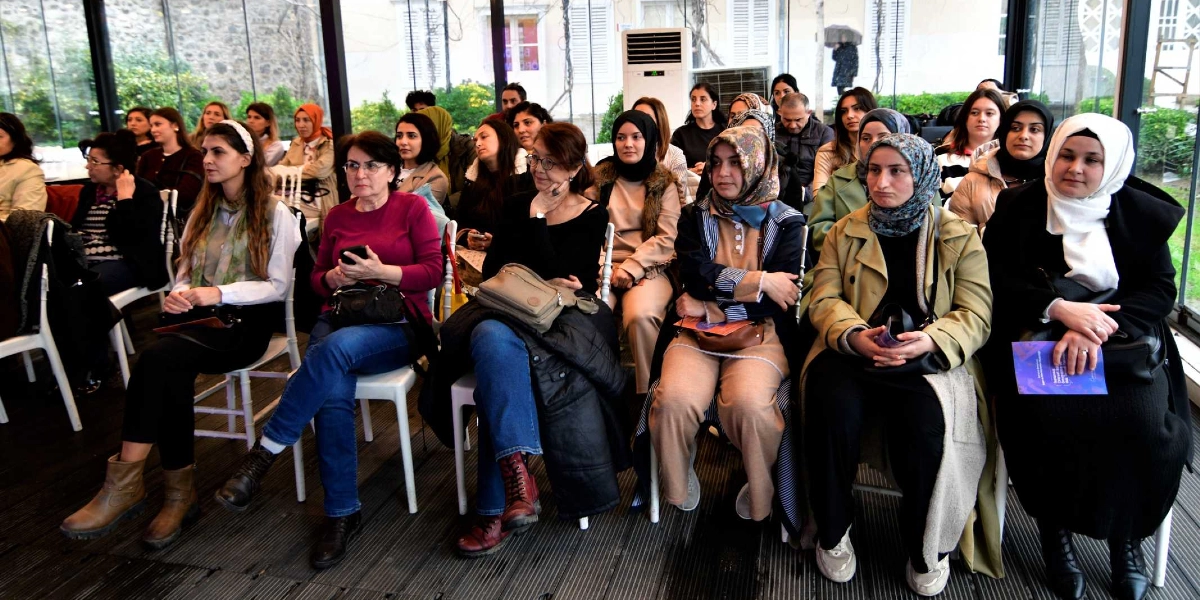 Cam duvarlarla çevrili aydınlık bir salonda, sandalyelerde oturmuş dikkatle bir konuşmayı dinleyen ağırlıklı olarak kadınlardan oluşan bir dinleyici grubu. Katılımcıların bir kısmı başörtülü, bir kısmı ise günlük kıyafetler içinde; bazıları ellerinde broşürler tutuyor. Arka planda ayakta duran bir klima ve dışarıdaki bahçeyi gösteren cam bölmeler yer alıyor.