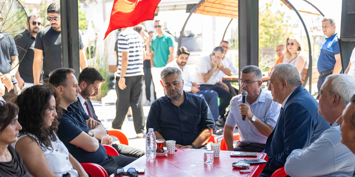 Açık havada düzenlenen bir anma toplantısında, kırmızı plastik bir masanın etrafında oturan bir grup insan yer alıyor. Masanın başında mikrofonla konuşma yapan gözlüklü bir adamın etrafında, onu dikkatle dinleyen katılımcılar bulunuyor. Arka planda ayakta duran diğer katılımcılar ve üst kısımda asılı duran Türk bayrağı görülüyor. Masanın üzerinde çay bardakları ve su şişeleri yer alıyor.