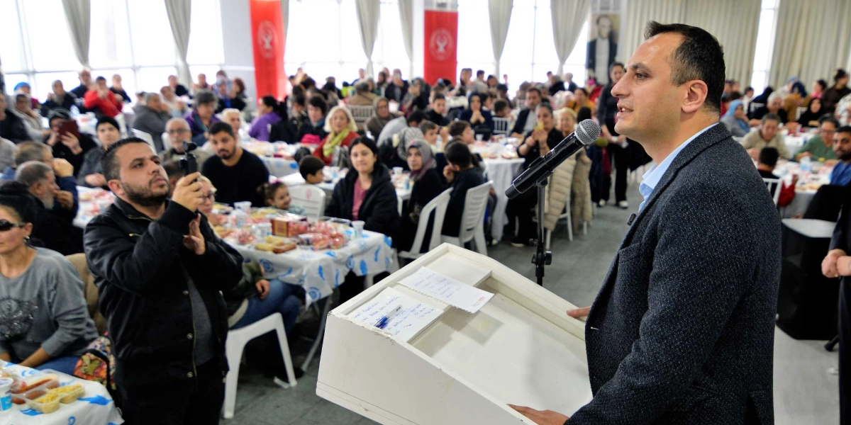 Bornova Belediye Başkanı Ömer Eşki, kalabalık bir katılımcı grubuna hitaben kürsüde konuşma yapıyor. Başkanın sağ tarafında, elindeki telefonla çekim yapan bir kişi bulunuyor. Arka planda ise masalarda oturan Bornovalı vatandaşlar yer alıyor.