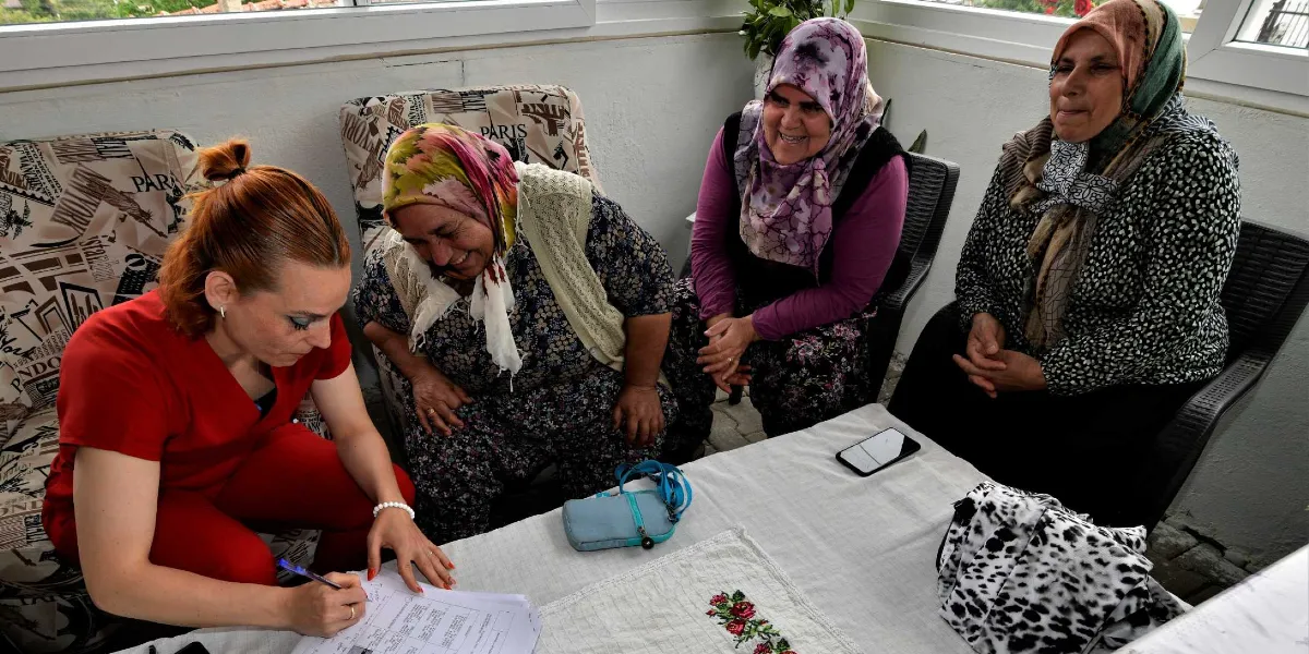 Kırmızı üniformalı bir sağlık görevlisi, beyaz örtülü bir masanın başında oturan üç kadına sağlık eğitimi vererek ellerindeki formları doldurmalarına yardımcı oluyor. Kadınlar başörtülü ve samimi bir ortamda oturuyorlar, masanın üzerinde bir cep telefonu ve küçük bir çanta bulunuyor.