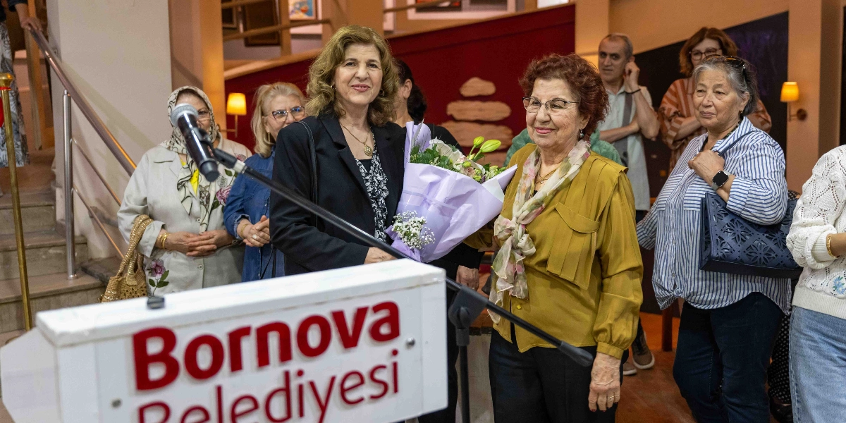 Bornova Belediyesi yazılı bir kürsünün önünde duran, elinde çiçek buketi tutan bir kadın ve yanında ona eşlik eden başka bir kadın ile arkalarında onları izleyen bir grup davetlinin bulunduğu bir sanat sergisi açılışı.