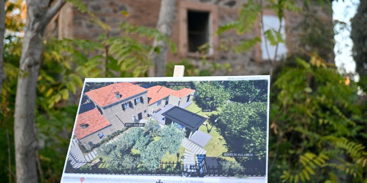 Açık havada, ağaçların önünde bir şövale üzerinde duran Girit Anı Evi projesine ait mimari yerleşim planı görseli. Üstten bakış açısıyla hazırlanan planda, taş dokulu ana bina, etkinlik salonu, kafe, tuvalet ve güvenlik kulübesinin yerleşimi işaretlenmiş. Planın arka planında ise projenin uygulanacağı tarihi taş yapının bulanık bir görüntüsü yer alıyor.