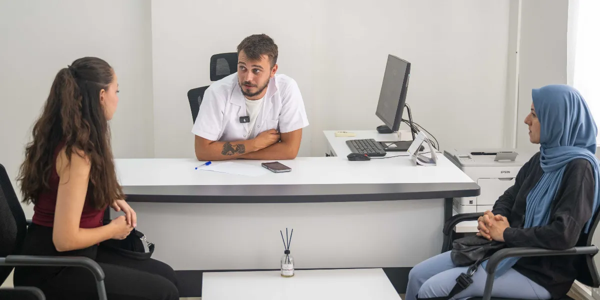 Beyaz önlüklü bir uzman, ofisindeki masasında oturan iki kadınla görüşme yapıyor. Uzman, masanın karşısında oturan ve biri başörtülü olan iki kadına dikkatle bakarak konuşuyor. Masanın üzerinde bir bilgisayar monitörü, klavye ve küçük bir oda kokusu bulunuyor.