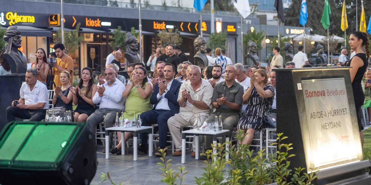 Bornova Belediyesi tarafından düzenlenen bir etkinlikte, katılımcılar açık havada sandalyelerde oturmuş alkışlıyor. Ön planda üzerinde 