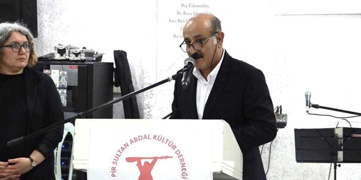 Pirsultan Abdal Kültür Derneği yazılı ve üzerinde derneğin logosu bulunan bir kürsüde konuşma yapan, gözlüklü ve bıyıklı bir erkek ile onun yanında duran gözlüklü bir kadın.