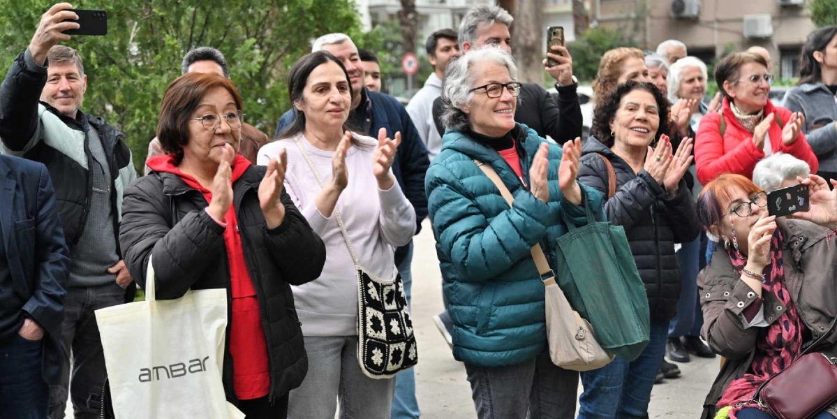 Açık havada bir araya gelmiş, gülümseyerek alkışlayan ve telefonlarıyla kayıt yapan bir grup insan. Ön planda, üzerinde 