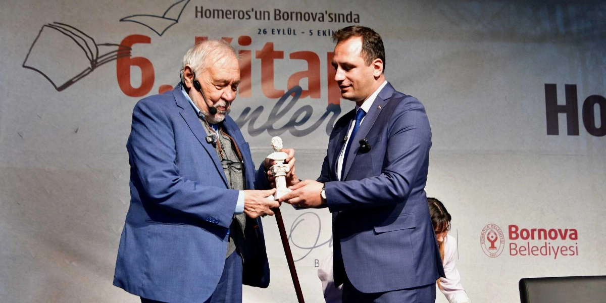 İlber Ortaylı, Bornova Belediyesi tarafından düzenlenen 6. Kitap Günleri etkinliğinde, kendisine takdim edilen Homeros büstünü Bornova Belediye Başkanı Mustafa İduğ'dan alırken görülüyor. Arka planda etkinliğin logosu ve tarihleri yer alıyor.