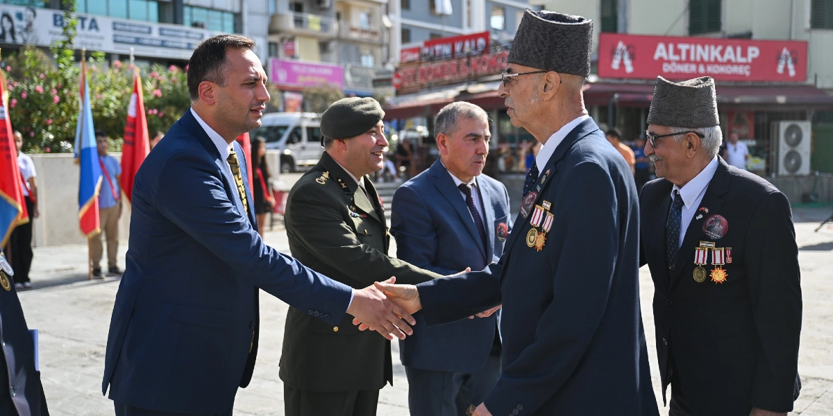 Bornova'daki 19 Eylül Gaziler Günü töreninde, lacivert takım elbiseli bir yetkili, üzerinde madalyaları ve kalpağı bulunan bir gazi ile tokalaşıyor. Yanlarında askeri üniformalı bir subay ve takım elbiseli başka bir gazi daha bulunuyor. Arka planda şehir meydanı, Türk bayrakları ve dükkan tabelaları yer alıyor.