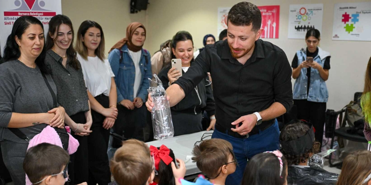 Bornova Belediyesi Altındağ Kültür Merkezi'nde düzenlenen bir etkinlikte, siyah gömlekli bir eğitmen çocuklara şeffaf bir su matarası gösteriyor. Eğitmenin etrafında onu ilgiyle izleyen çocuklar ve onları gözlemleyen yetişkinler bulunuyor. Arka planda duvarda Bornova Belediyesi'ne ait bilgilendirici afişler yer alıyor.