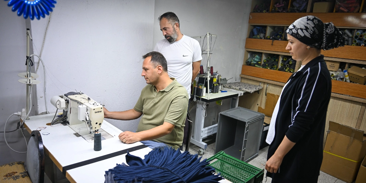 Küçük bir atölyede, zeytin yeşili tişört giyen bir adam Brother marka dikiş makinesinde çalışırken, yanında beyaz tişörtlü bir adam ve başında desenli bir eşarp bulunan kadın ayakta durarak onu izliyor. Arka planda raflara dizilmiş çok sayıda iplik makarası ve karton kutular bulunuyor.