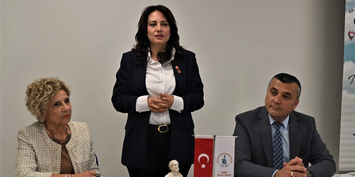 Beyaz bir arka plan önünde, ortada ayakta duran koyu renk ceketli ve beyaz gömlekli bir kadın konuşma yaparken, yanında oturan kıvırcık saçlı bir kadın ve gri takım elbiseli bir erkek onu dinliyor. Masanın üzerinde küçük bir Türk bayrağı ve yanında İzmir Bornova Belediyesi'ne ait olduğu anlaşılan bir flama ile küçük bir büst yer alıyor.