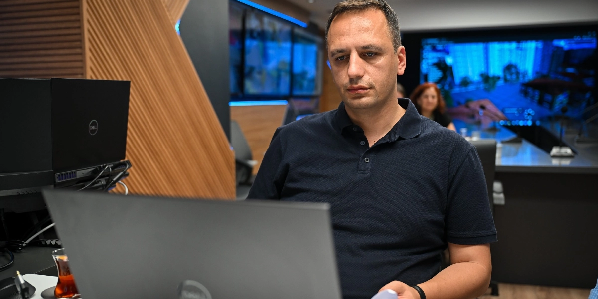 Koyu lacivert polo yaka tişört giymiş bir erkek, modern bir ofis ortamında dizüstü bilgisayarına odaklanmış şekilde çalışıyor. Arka planda büyük ekranlı bir kontrol merkezi veya toplantı odası düzeni görülüyor.