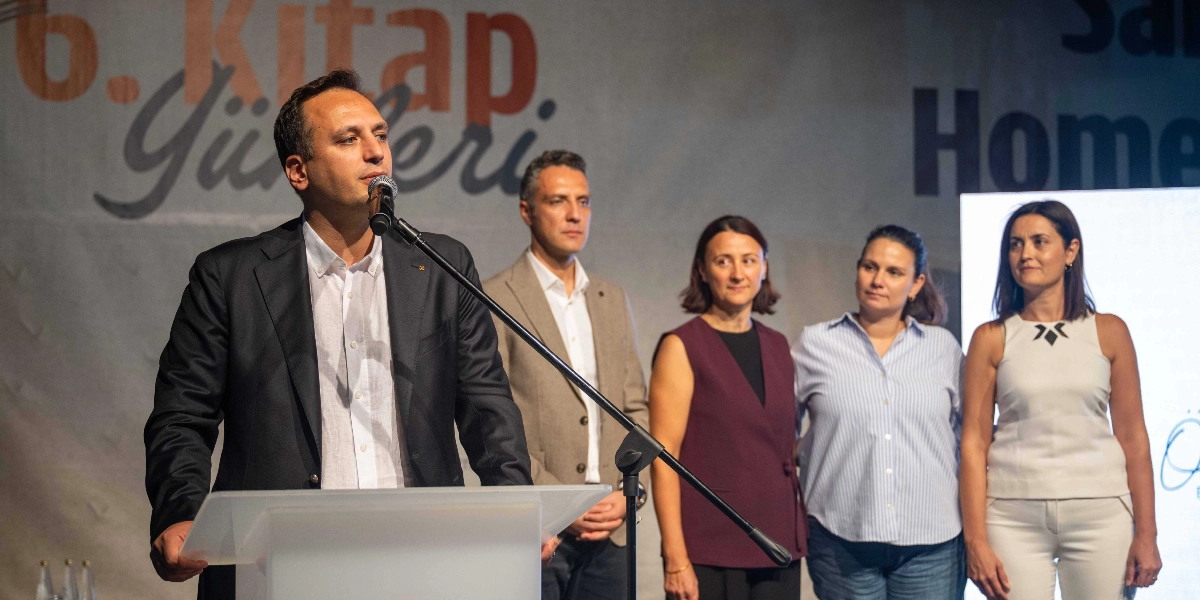 Bornova 6. Kitap Günleri etkinliğinde, kürsüde konuşma yapan bir erkek ve arkasında yan yana duran dört kişilik bir grup bulunuyor. Konuşmacı koyu renkli bir ceket ve beyaz gömlek giymiş, mikrofon başında izleyicilere hitap ediyor. Arka planda ise üzerinde 