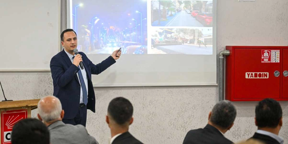 Koyu renk takım elbiseli bir erkek, elinde mikrofonla bir sunum yaparken arkasındaki projeksiyon perdesini işaret ediyor. Perdede, gece aydınlatılmış bir cadde ile asfalt çalışması yapılan bir sokağın fotoğrafları yer alıyor. Konuşmacının sol tarafında kürsü üzerinde CHP logosu, sağ tarafta ise duvara monte edilmiş kırmızı bir yangın dolabı bulunuyor. İzleyiciler, konuşmacıya arkaları dönük şekilde sunumu takip ediyor.