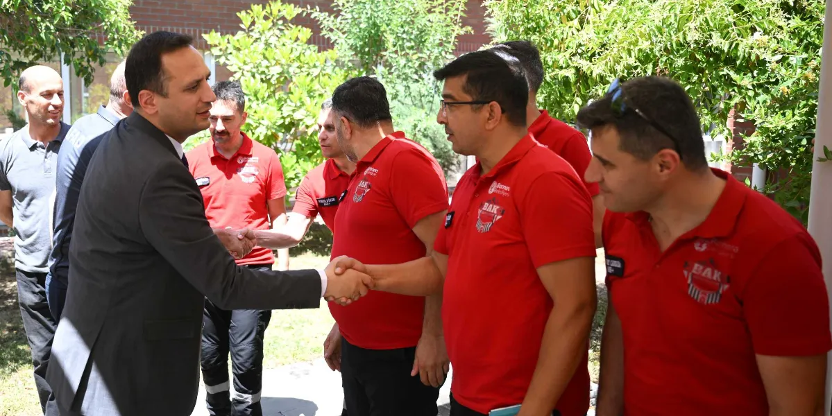 Takım elbiseli bir yetkili, üzerinde Bornova Belediyesi Arama Kurtarma (BAK) logolu kırmızı tişörtler bulunan arama kurtarma ekibi üyeleriyle açık havada tokalaşarak tebrik ediyor.