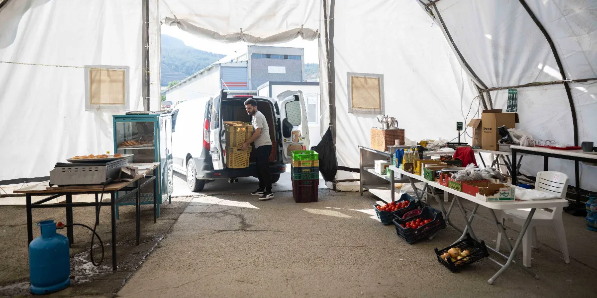 Beyaz bir çadırın içinde, bir genç adam beyaz bir hafif ticari aracın arka kapısından sarı plastik kasalar indiriyor. Çadırın içinde, üzerinde domates ve soğan gibi sebzelerin bulunduğu kasaların dizili olduğu masalar, bir mutfak tezgahı, tüp ve çeşitli gıda malzemeleri yer alıyor. Arka planda ise dışarıdaki açık alan ve uzaktaki binalar görünüyor.