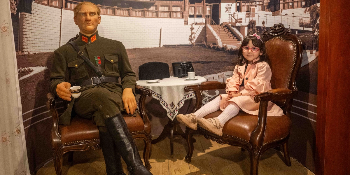 Belkahve Ata Anı Evi'nde, askeri üniformalı Mustafa Kemal Atatürk'ün balmumu heykeli ile yanındaki koltukta oturan küçük bir kız çocuğu. Atatürk, elinde bir fincan kahveyle deri bir koltukta otururken, yanında dantel örtülü küçük bir masada dürbün ve başka bir fincan bulunuyor. Arka planda, üzerinde 