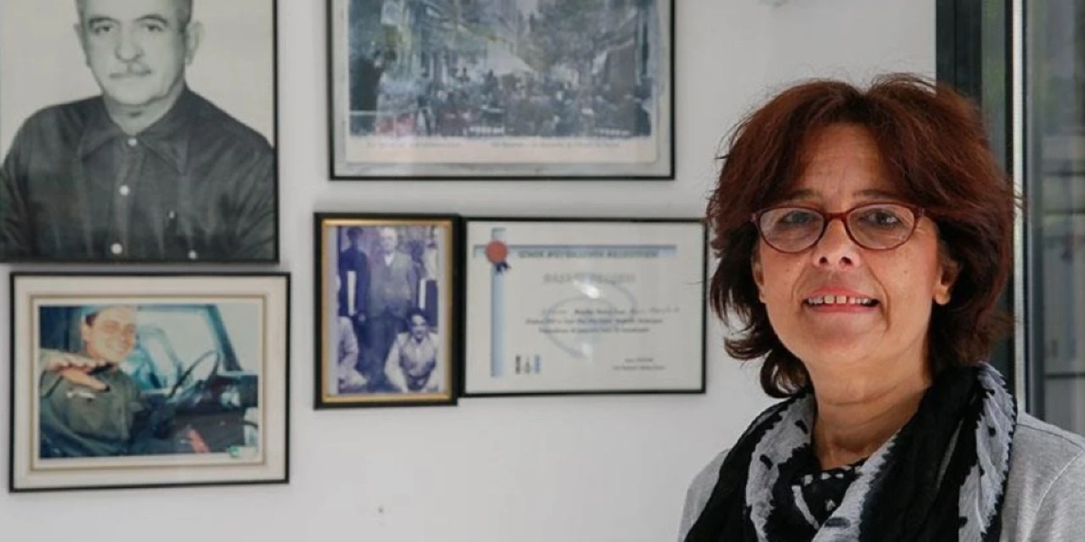 Kızıl saçlı, gözlüklü ve boynunda desenli bir şal bulunan orta yaşlı bir kadın, beyaz bir duvarın önünde duruyor. Kadının arkasındaki duvarda, siyah beyaz bir erkek portresi, eski bir kent meydanını gösteren bir fotoğraf, başka bir eski fotoğraf ve İzmir Bornova Belediyesi'ne ait bir başarı belgesi çerçevelenmiş şekilde asılı duruyor.
